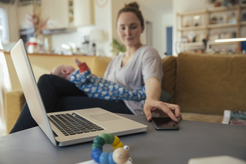 Mother with newborn baby at home taking cell phone from table stock photo