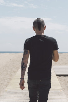 Rückenansicht eines jungen Mannes mit Irokesenschnitt und Tätowierungen, der auf der Promenade zum Strand läuft - SKCF00303