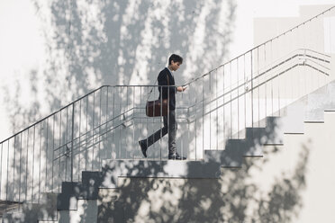 Junger Mann im Anzug geht auf einer Treppe und schaut auf sein Handy - SKCF00306