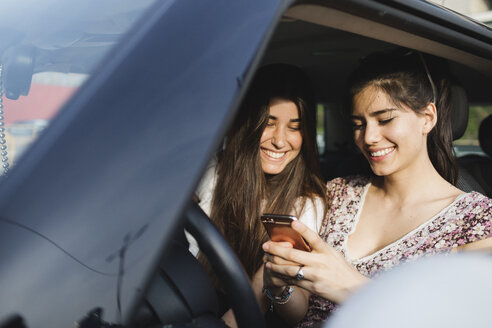 Zwei glückliche junge Frauen schauen in einem Auto auf ihr Handy - MRAF00192