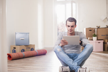 Junger Mann in neuer Wohnung sitzt mit Tablet auf dem Boden - UUF10705