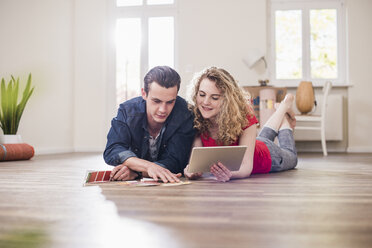Junges Paar in neuem Haus, auf dem Boden liegend, mit einem Tablet, das ein Farbmuster auswählt - UUF10741
