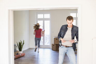 Young man in new home using tablet with woman in background carrying cardboard box - UUF10745