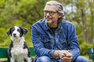 Porträt eines Mannes mit grauem Haar und Bart, der neben seinem Hund auf einer Bank sitzt - TCF05412