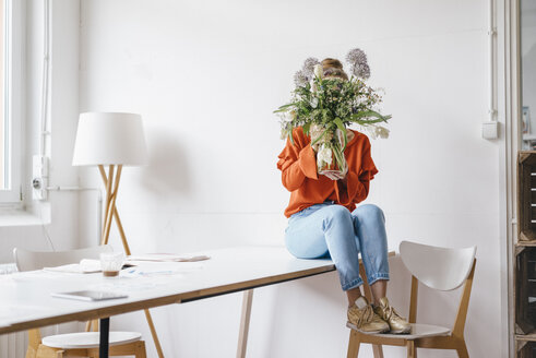 Young woman sitting on table holding flower vase in front of her face - KNSF01459