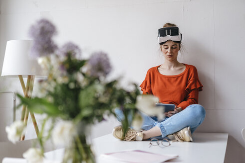 Young woman sitting on table using tablet wearing VR glasses - KNSF01514