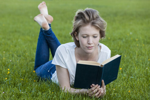 Barefoot young woman lying on a meadow reading a book - TCF05427