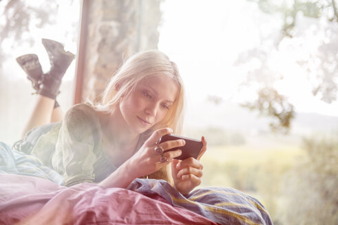 Porträt einer jungen Frau, die auf dem Bett liegt und auf ihr Handy schaut - ZOCF00411