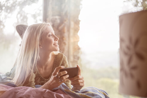 Porträt einer lächelnden jungen Frau, die mit ihrem Handy auf dem Bett liegt und durch das Fenster schaut - ZOCF00412