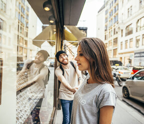 Junge Frau schaut fasziniert in ein Schaufenster, ihr Freund versucht, sie wegzuziehen - AIF00430