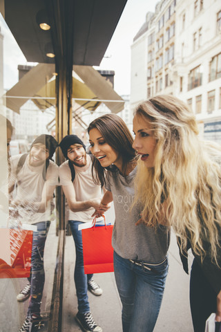 Junge Frauen schauen fasziniert in ein Schaufenster, ihr Freund versucht, sie wegzuziehen, lizenzfreies Stockfoto