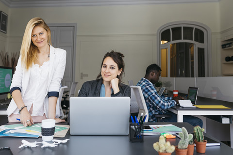 Junge Geschäftsfrauen arbeiten zusammen, benutzen einen Laptop, ein Kollege sitzt im Hintergrund, lizenzfreies Stockfoto