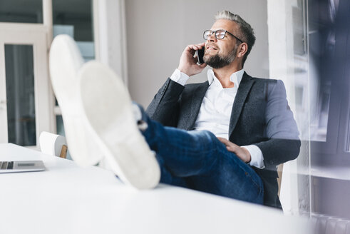 Businessman sitting phoning in office with feet on desk - JOSF01016