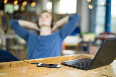 Laptop und Smartphone auf einem Tisch mit einem entspannten jungen Mann, der im Hintergrund Musik hört - JOSF01036