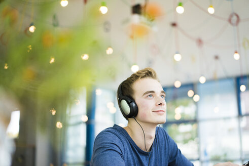 Porträt eines lächelnden jungen Mannes, der in einem Café mit Kopfhörern Musik hört - JOSF01041