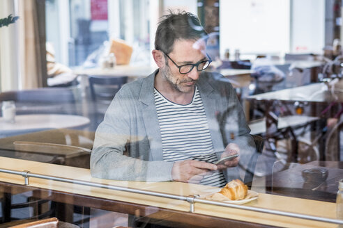 Älterer Mann sitzt hinter der Fensterscheibe eines Cafés und schaut auf sein Handy - FMKF04154