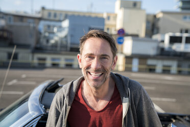 Porträt eines lachenden reifen Mannes mit Bartstoppeln vor seinem Sportwagen - FMKF04165