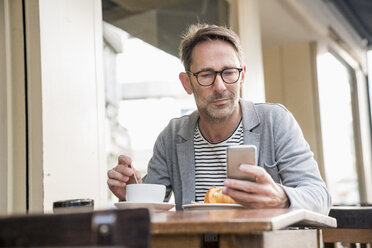 Portrait of mature man sitting at sidewalk cafe looking at cell phone - FMKF04169