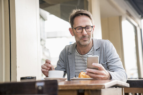 Porträt eines reifen Mannes, der in einem Straßencafé sitzt und auf sein Mobiltelefon schaut - FMKF04169