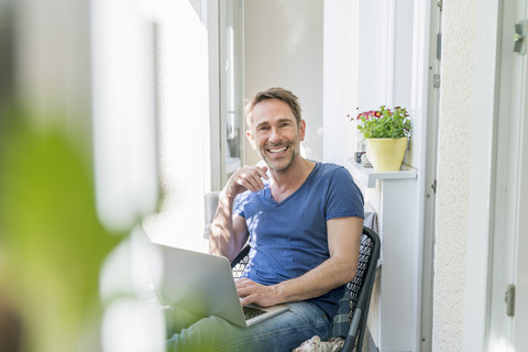 Porträt eines lächelnden reifen Mannes, der mit einem Laptop auf einem Balkon sitzt, lizenzfreies Stockfoto