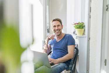 Porträt eines lächelnden reifen Mannes, der mit einem Laptop auf einem Balkon sitzt - FMKF04175
