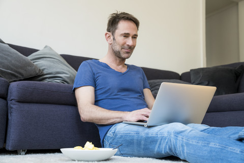 Portrait of smiling man sitting on the floor in the living room using laptop stock photo