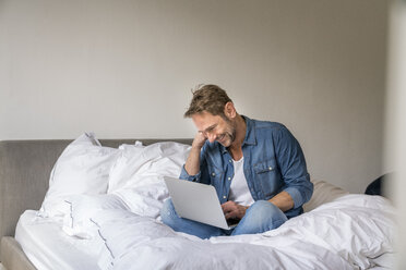 Smiling mature man sitting on his bed using laptop - FMKF04183