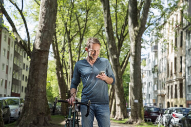 Smiling mature man with bicycle in the city looking at cell phone - FMKF04188