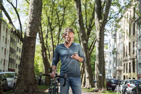Lächelnder reifer Mann mit Fahrrad in der Stadt, der auf sein Handy schaut - FMKF04188