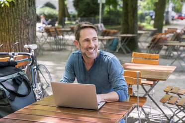 Porträt eines lächelnden reifen Mannes mit Laptop im Biergarten - FMKF04190