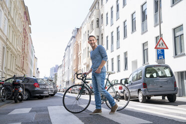 Porträt eines lächelnden reifen Mannes mit Fahrrad beim Überqueren der Straße - FMKF04193