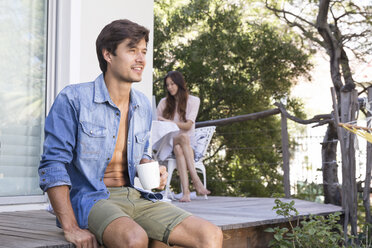 Man sitting on terrace with cup of coffee and woman in background - WESTF23168