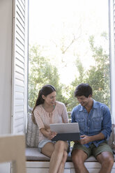 Smiling young couple sitting on windowsill sharing tablet - WESTF23197