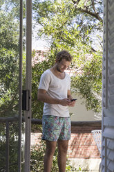 Junger Mann in Badehose und T-Shirt telefoniert mit Handy - WESTF23244