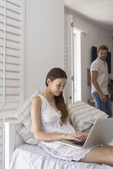 Junge Frau benutzt Laptop im Schlafzimmer mit Mann im Hintergrund - WESTF23247
