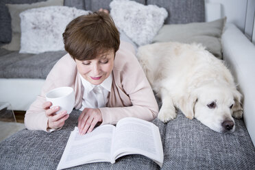 Ältere Frau auf der Couch liegend, Buch lesend mit Hund an ihrer Seite - WESTF23278