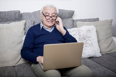 Älterer Mann sitzt auf der Couch und benutzt Laptop und Smartphone - WESTF23299