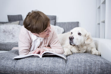 Senior woman lying on couch, reading book with dog by her side - WESTF23311