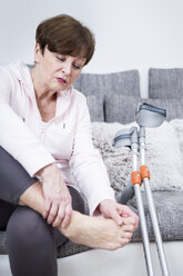 Ältere Frau mit Krücken sitzt auf der Couch und überprüft ihren Knöchel - WESTF23338