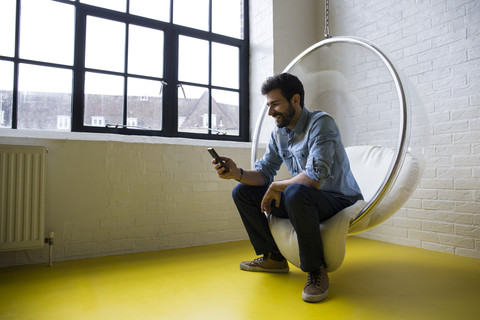 Lächelnder Mann sitzt auf einer Schaukel in seinem Loft und schaut auf sein Handy, lizenzfreies Stockfoto