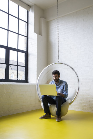 Lächelnder Mann sitzt auf einer Schaukel in seinem Loft und benutzt einen Laptop, lizenzfreies Stockfoto