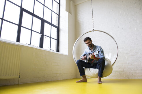 Junger Mann sitzt auf einer Schaukel auf seinem Dachboden und spielt Gitarre - ABZF02047