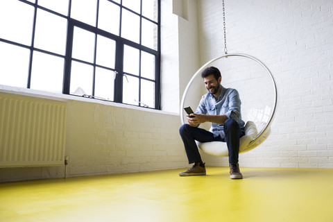 Junger Mann sitzt auf einer Schaukel in seinem Loft und schaut auf sein Handy, lizenzfreies Stockfoto