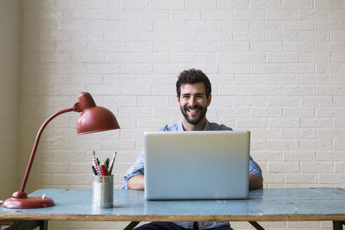 Portrait of happy young man sitting at desk working with laptop - ABZF02068