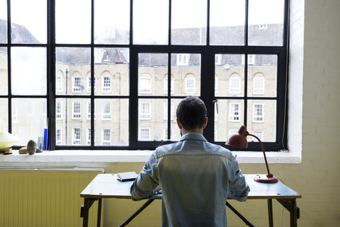 Rückenansicht eines jungen Mannes, der in seinem Loft am Schreibtisch sitzt und mit seinem Laptop arbeitet, lizenzfreies Stockfoto
