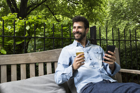 Porträt eines lächelnden jungen Mannes mit Mobiltelefon und einem Glas Bier, der sich im Garten entspannt - ABZF02077