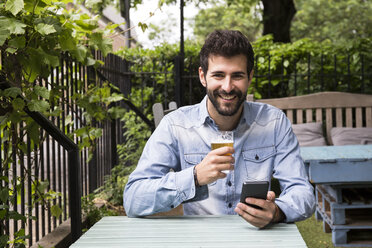 Porträt eines lächelnden jungen Mannes mit Mobiltelefon und einem Glas Bier, der sich im Garten entspannt - ABZF02078