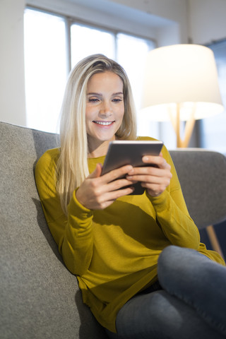 Porträt einer lächelnden Frau, die auf einer Couch sitzt und ein Mini-Tablet benutzt, lizenzfreies Stockfoto