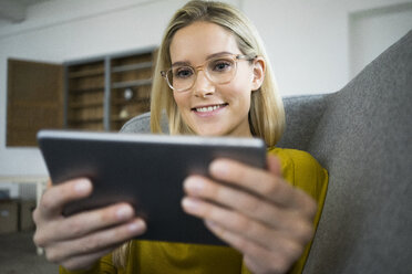 Porträt einer lächelnden Frau mit Brille, die auf einer Couch sitzt und ein Mini-Tablet benutzt - JOSF01068