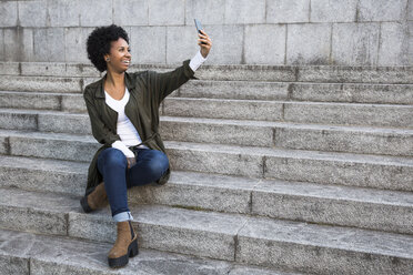 Lächelnde junge Frau sitzt auf einer Treppe und macht ein Selfie mit ihrem Smartphone - ABZF02089